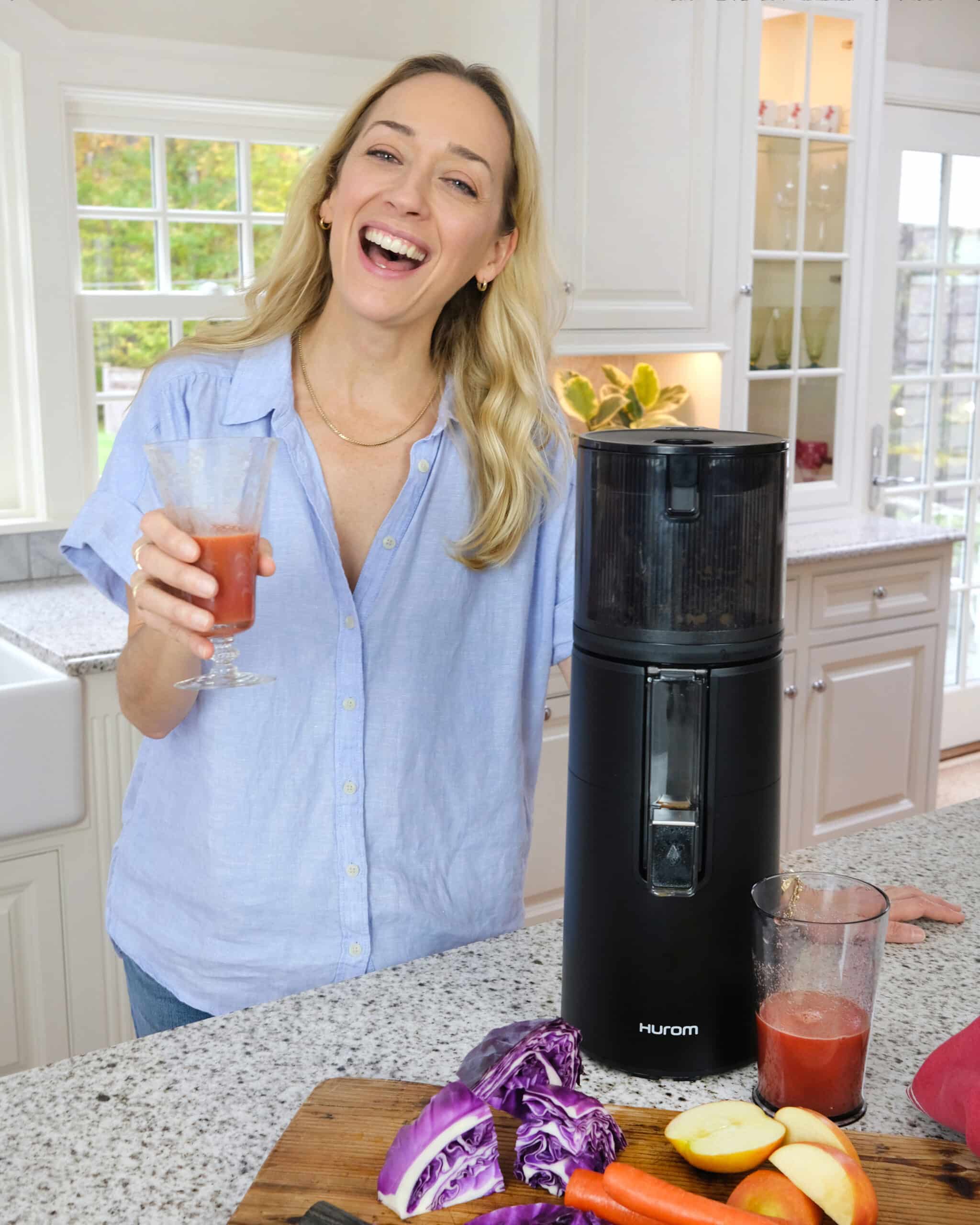 Kris Carr standing beside a juicer holding a glass of juice.